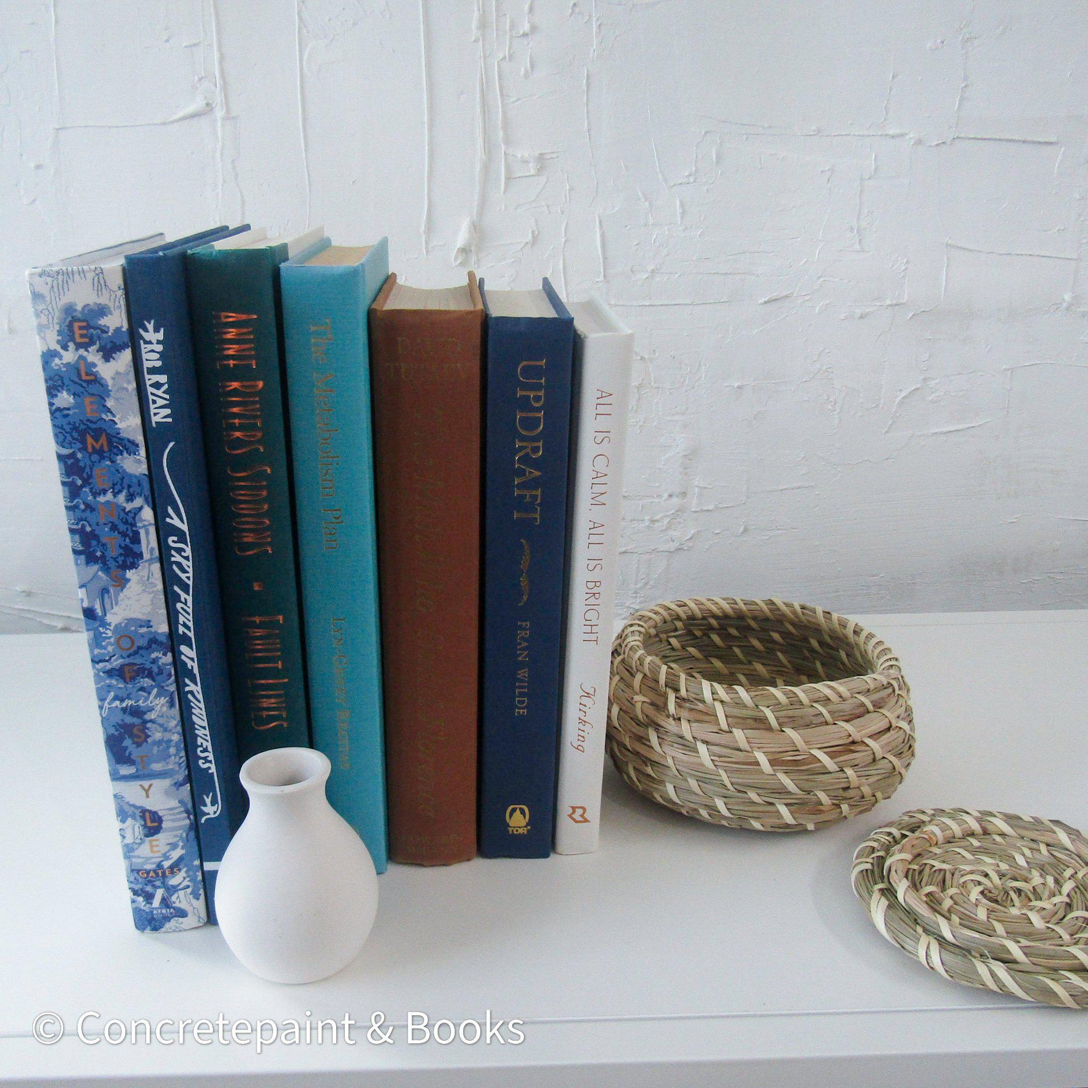 Real hardcover books with ikea seagrass basket and small white vase staged as home décor. Blue, white, green and brown hardcover books.