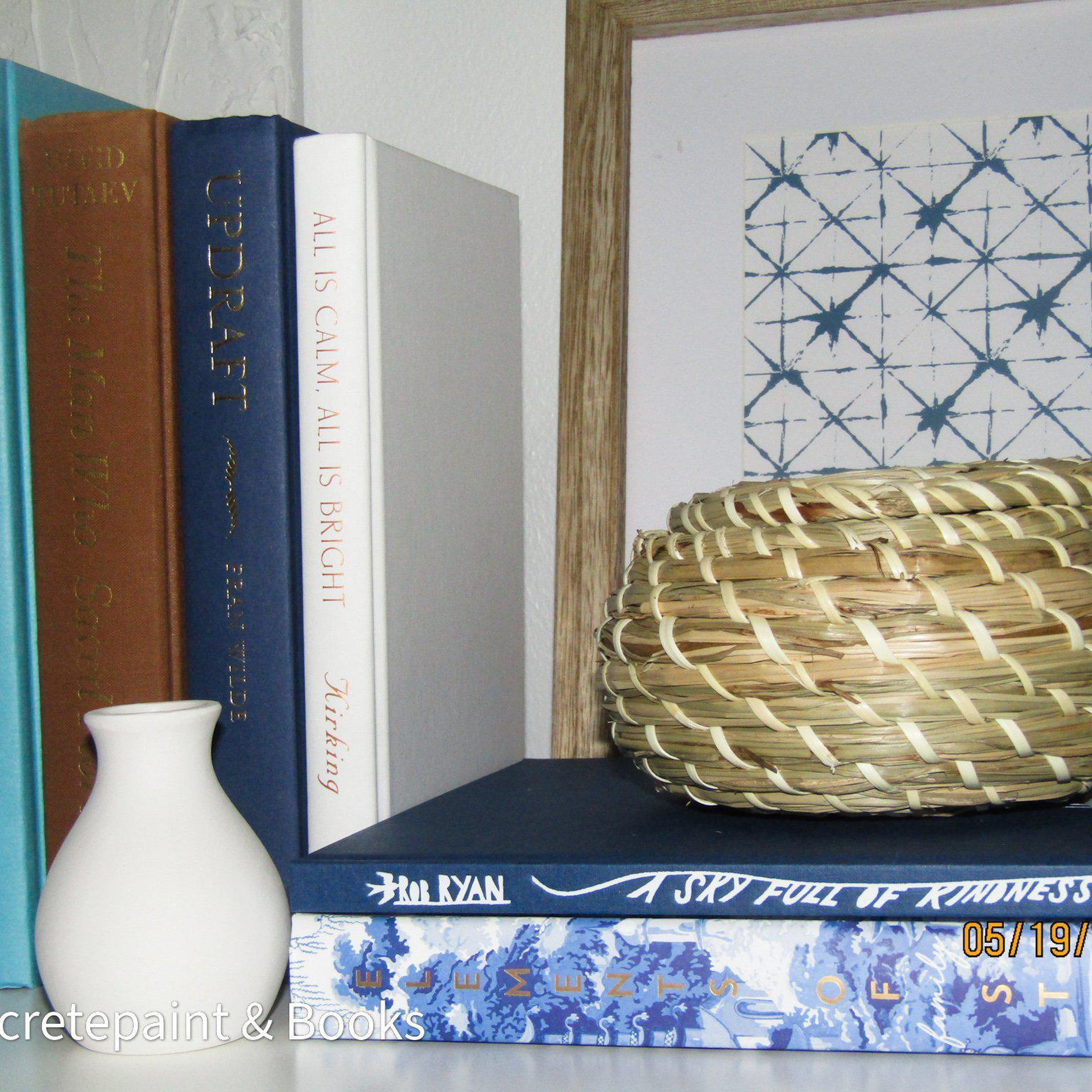 Real hardcover books with ikea seagrass basket and small white vase staged as home décor. Blue, white, green and brown hardcover books.